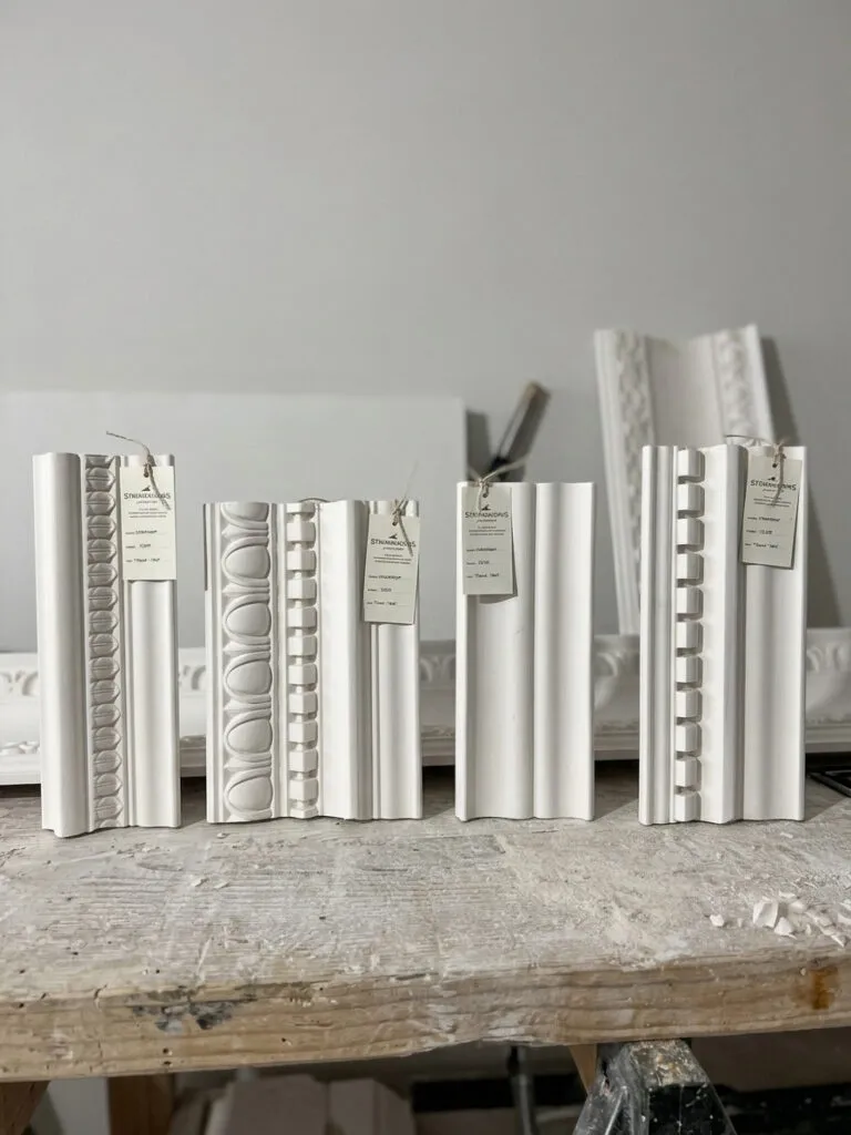 A selection of four traditional white plaster crown moulding and cornice samples standing upright on a dusty wooden workshop bench. The detailed architectural profiles feature classical motifs like dentil blocks and egg-and-dart borders, complete with hanging paper spec tags.