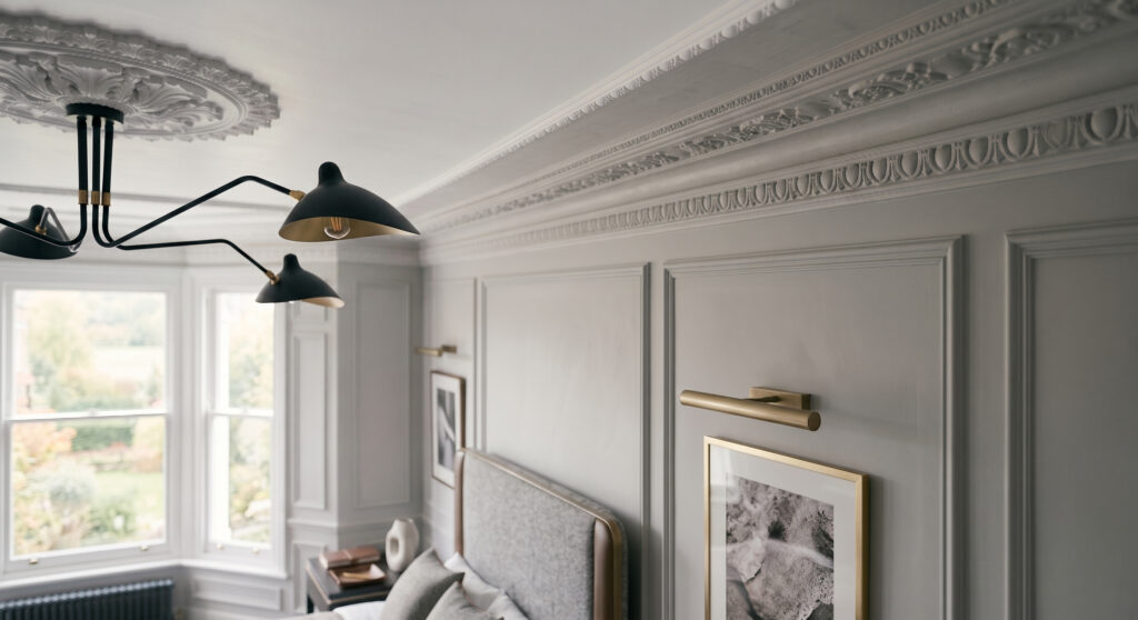 A dramatic, low-angle shot of a sophisticated master bedroom featuring muted gray-panelling on the walls. The ceiling showcases an intricate, classical plaster ceiling rose paired with a contrasting, modern black multi-arm spider chandelier with brass accents. A bespoke bed with a textured gray fabric headboard and a dark minimalist nightstand with brass linear picture lights are visible in the foreground.