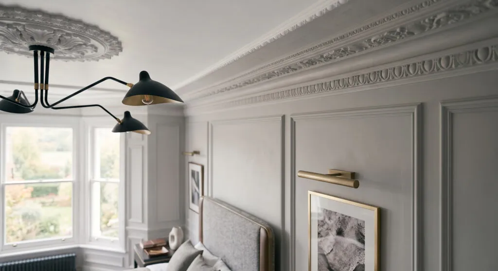A dramatic, low-angle shot of a sophisticated master bedroom featuring muted gray-panelling on the walls. The ceiling showcases an intricate, classical plaster ceiling rose paired with a contrasting, modern black multi-arm spider chandelier with brass accents. A bespoke bed with a textured gray fabric headboard and a dark minimalist nightstand with brass linear picture lights are visible in the foreground.