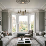 A perfectly balanced, wide-angle shot of an elegant London living room in Hampstead. Two identical Minotti-style sofas in textured gray upholstery sit opposite each other on a thick wool rug. A tiered dark wood and chrome coffee table centers the room under a traditional brass branch chandelier. A classic three-sided bay window with crisp white-painted frames opens up to a verdant park view, perfectly framed by sophisticated gray wall panel moulding and historical cornicing.
