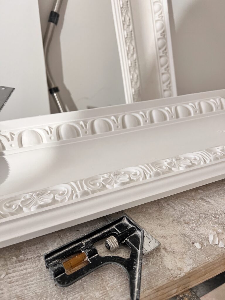 A close-up detail shot of a pristine white plaster crown moulding profile resting on a wooden workshop bench covered in fine plaster dust. The moulding features classic egg-and-dart and acanthus leaf decorative details. A black and silver metal combination adjustable square ruler sits sharply in the foreground.