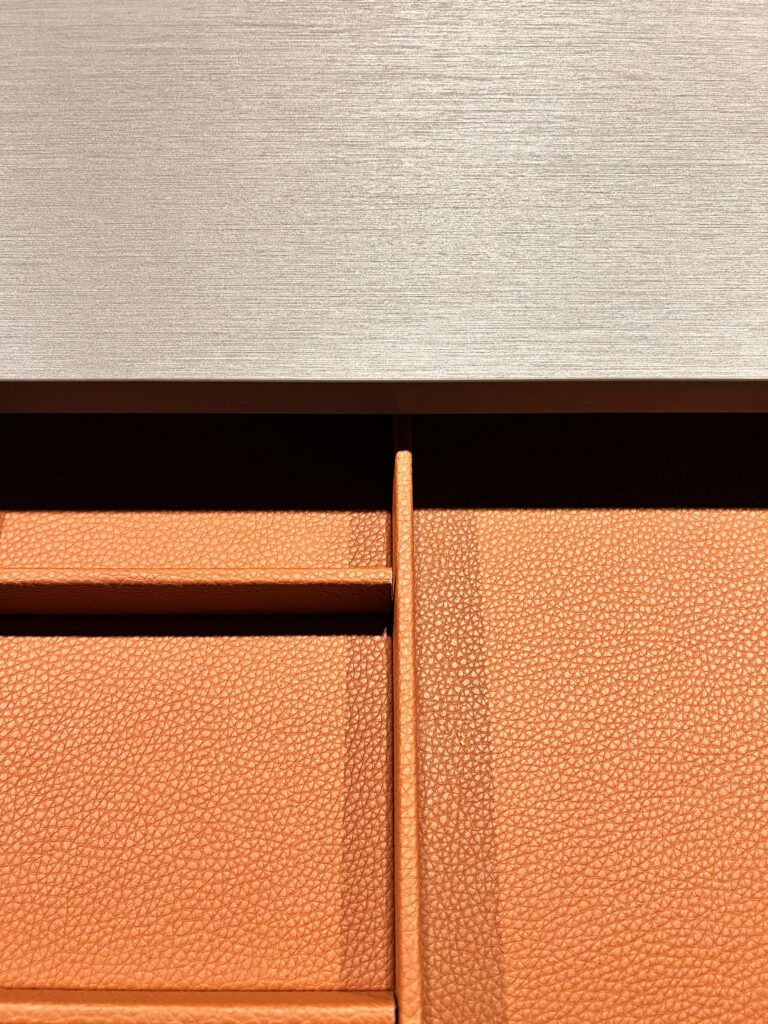 Macro detail of custom Italian wardrobe drawer dividers lined in pebbled terracotta leather beneath a metallic brushed counter surface