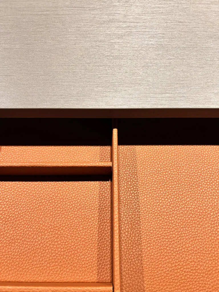 Macro detail of custom Italian wardrobe drawer dividers lined in pebbled terracotta leather beneath a metallic brushed counter surface