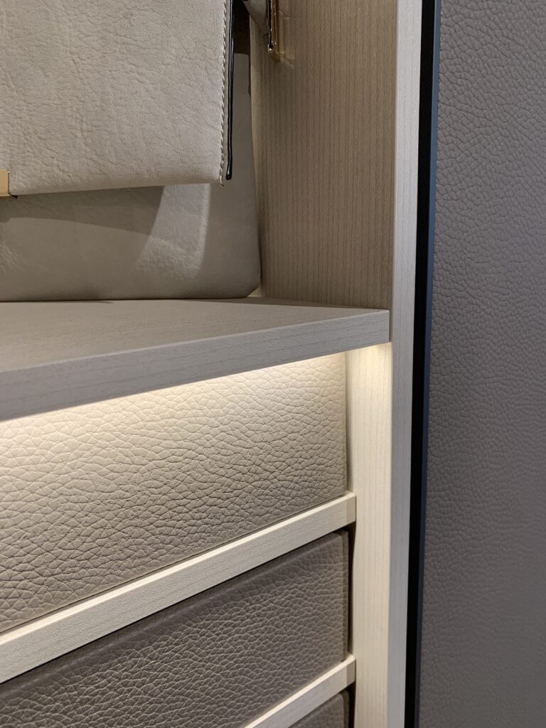 Close-up detail of bespoke Italian wardrobe interior featuring pebbled leather drawer fronts, light oak shelving, and integrated warm micro-LED strip lighting.