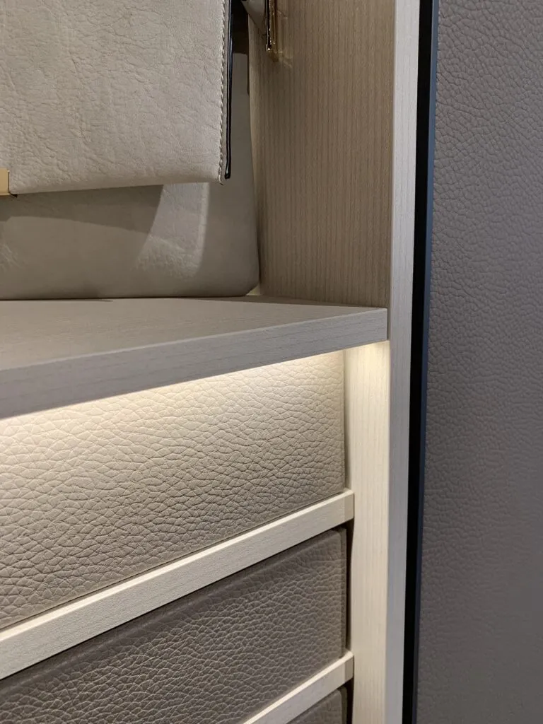 Close-up detail of bespoke Italian wardrobe interior featuring pebbled leather drawer fronts, light oak shelving, and integrated warm micro-LED strip lighting.