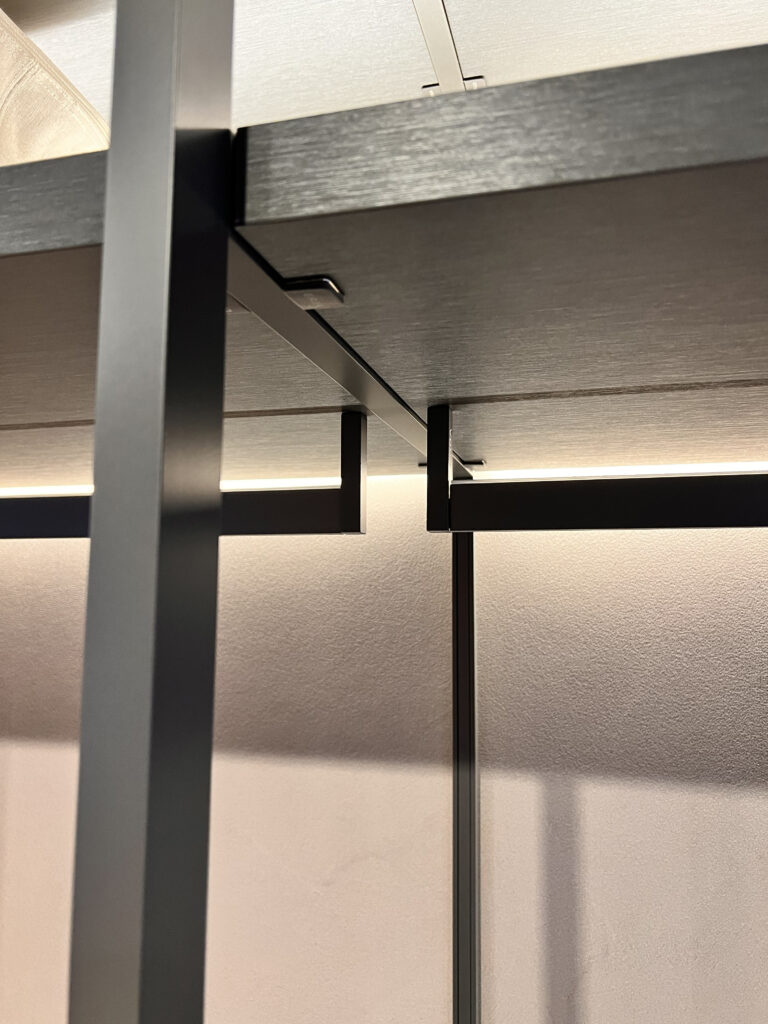 Close-up architectural detail of a bespoke Italian wardrobe showing a matte black metal structural frame, dark timber shelving, and an integrated recessed LED hanging rail.