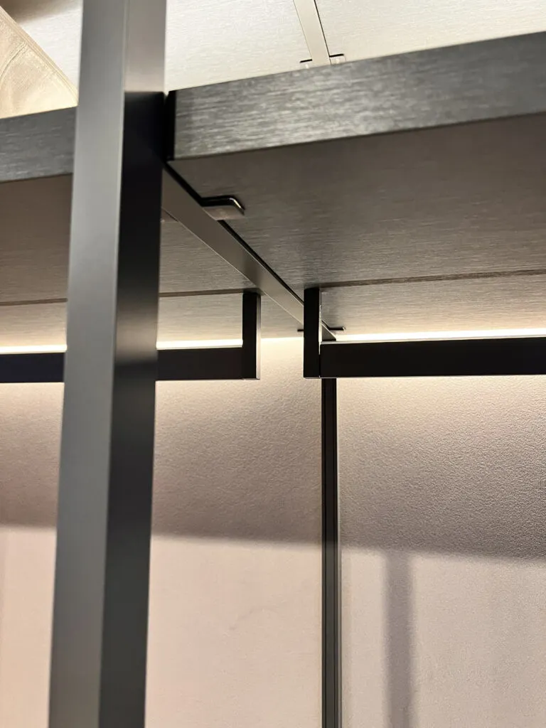 Close-up architectural detail of a bespoke Italian wardrobe showing a matte black metal structural frame, dark timber shelving, and an integrated recessed LED hanging rail.