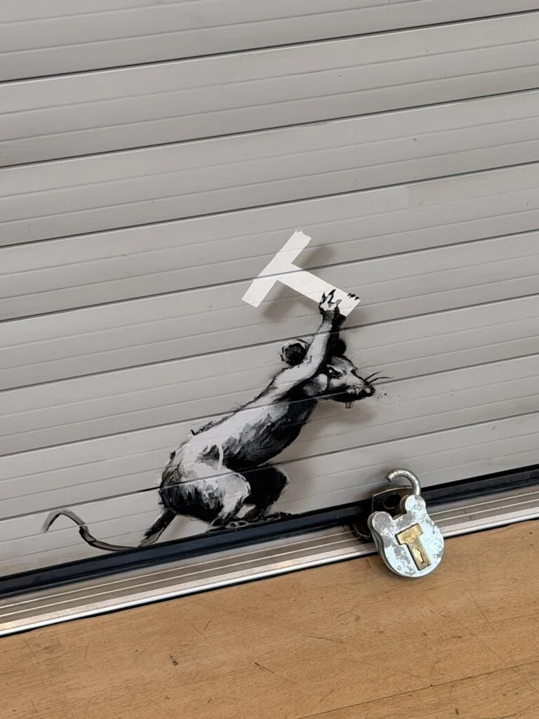 Detail view of Banksy stenciled black rat holding the letter T as a chisel next to an open brass padlock on airport shutter