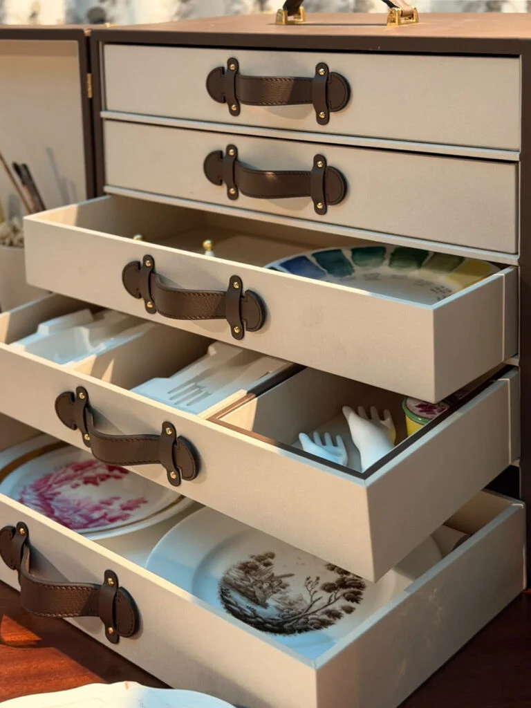 Open multi-tiered luxury collector chest with stitched leather pull handles and custom-molded felt compartments storing fine porcelain plates