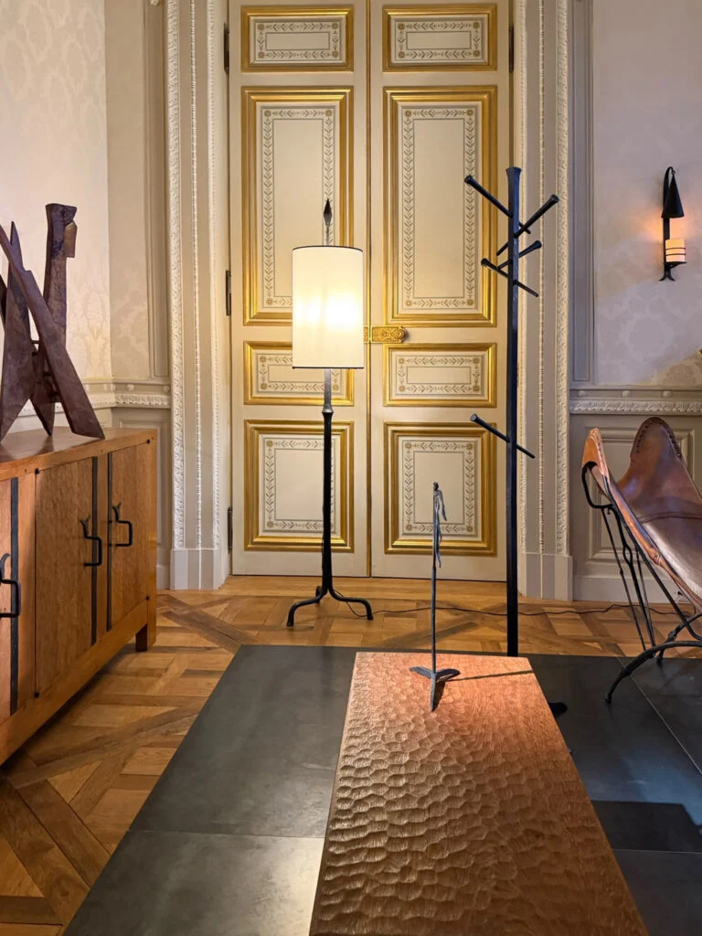 Hand-sculpted textured timber bench, brutalist forged iron floor lamp, and coat rack standing before gilded classical double doors