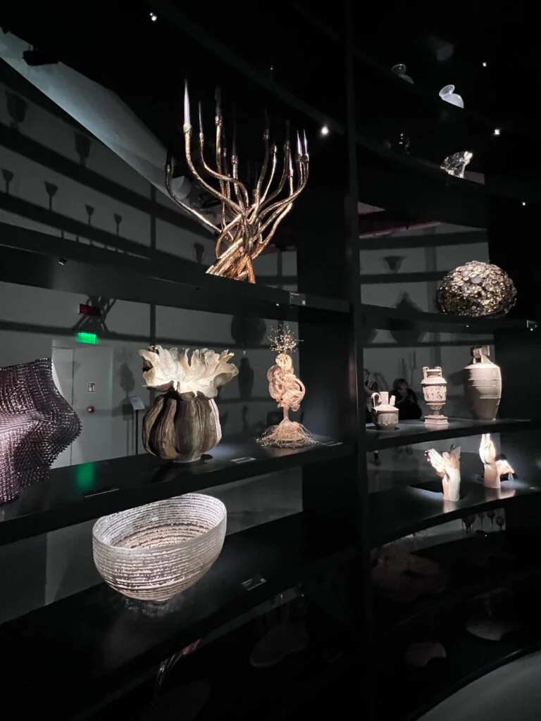 A curated dark wood exhibition shelf display at Homo Faber, showcasing a large, sculpted golden branched candelabra chandelier above, a textured glass bowl below, and various bespoke ceramic and metal objects, including an embossed pot and sculptural hands.