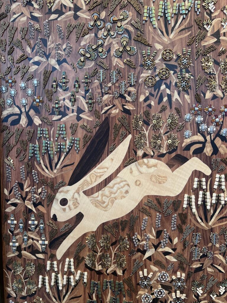 Close-up of an intricate decorative wood marquetry panel at Homo Faber depicting a running hare inlaid with figured timber grain, surrounded by detailed micro-beaded metallic embroidery and floral motifs. Perfect for luxury interior designers in London
