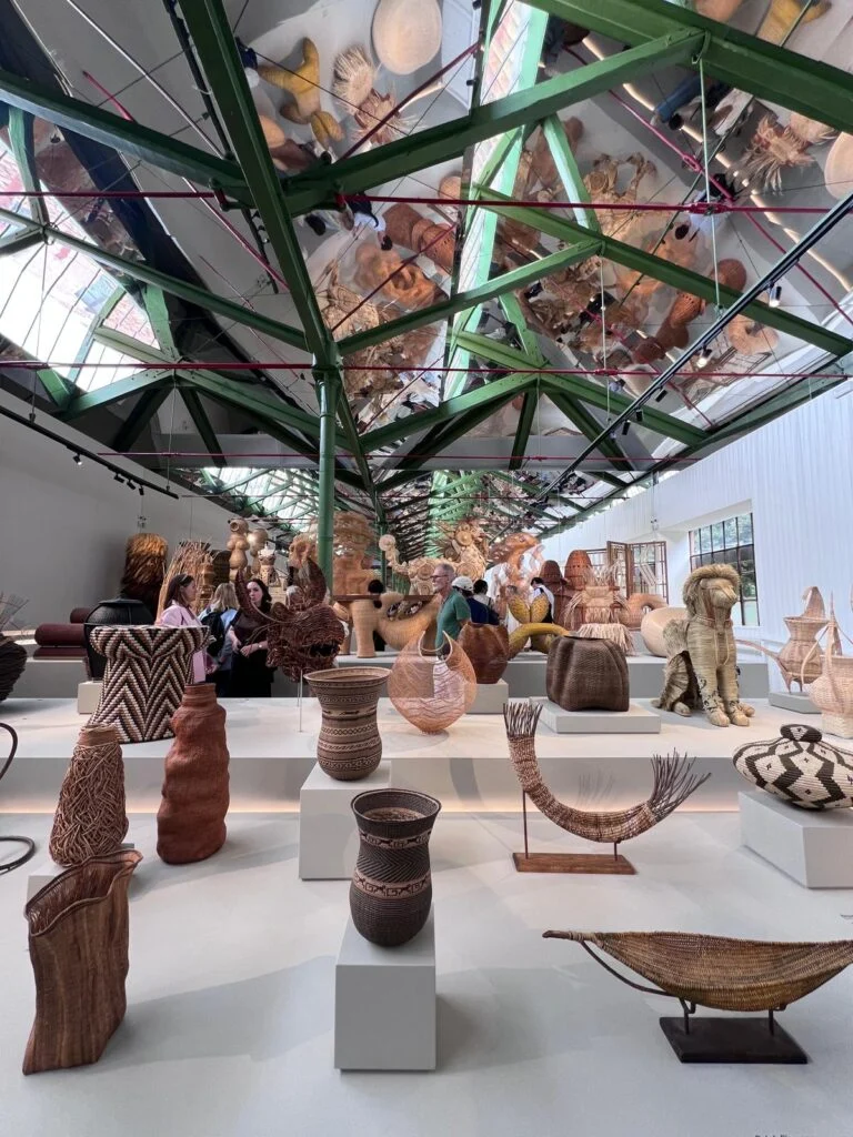 Exhibition hall at Homo Faber Venice under exposed green architectural steel trusses with mirrored ceiling panels, featuring an array of bespoke handcrafted woven fiber vessels, textural baskets, and organic zoomorphic sculptures.
