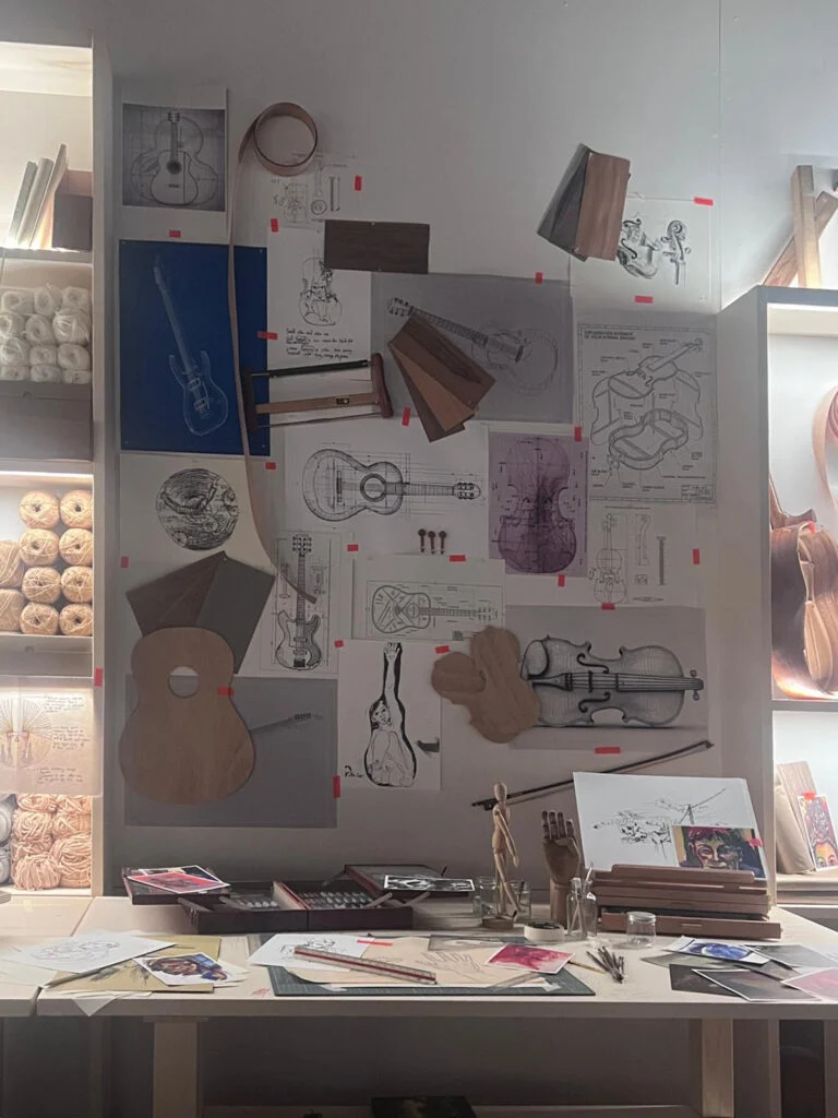 Workbench and inspiration mood board of a master luthier at Homo Faber Venice, featuring technical pencil sketches, violin and guitar blueprints, wood veneer swatches, timber templates, and artisan tools.