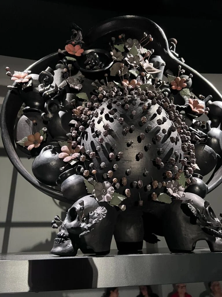 Close-up view of a complex, hand-sculpted Mexican ceramic 'Tree of Life' style sculpture at Homo Faber, featuring multiple dark burnished clay pots, tiny sculpted skulls on pins, and decorative pink flowers, on display.