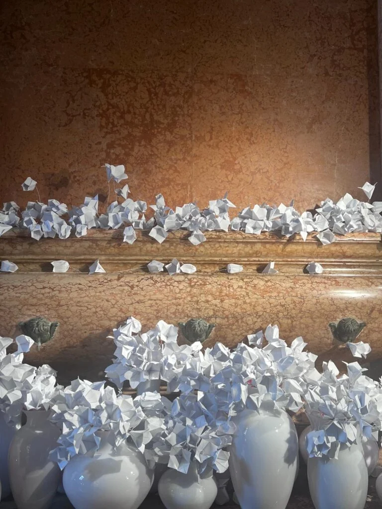 culptural white folded origami paper flowers displayed inside glossy ceramic vases and cascading across an antique veined breccia marble fountain basin at Homo Faber Venice.