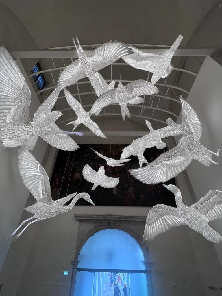 Impressive architectural ceiling installation at Homo Faber Venice featuring luminous, feather-textured translucent glass bird sculptures suspended in flight within a historic vaulted hall