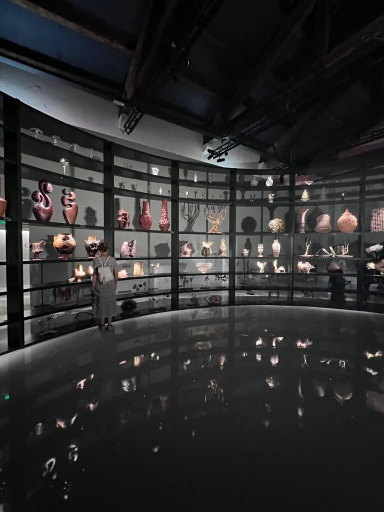 Wide panoramic photograph of a large, curved, dark metal exhibition hall at Homo Faber, Venice, filled with countless unique artisan ceramic, glass, and metal objects on multiple tiered shelves. A person is seen viewing the display, which reflects in the polished floor.