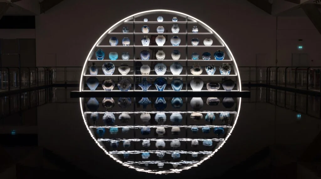 Symmetrical view of a large semi-circular illuminated display shelf with multiple tiers of unique, handcrafted blue and white detailed ceramic vases, perfectly reflected in dark, still water at Homo Faber exhibition in Venice.