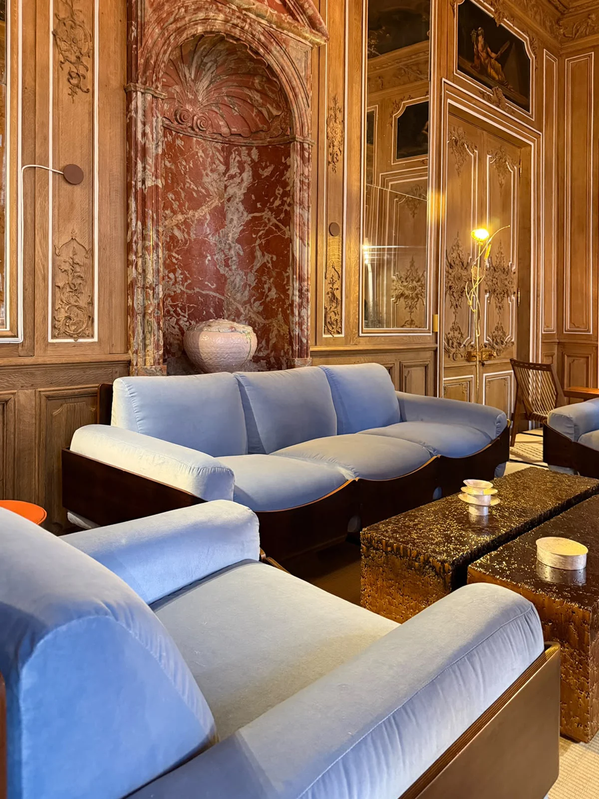 Powder blue velvet curved modular sofa and brutalist textured bronze coffee tables framed by an antique Rouge de Rance marble niche